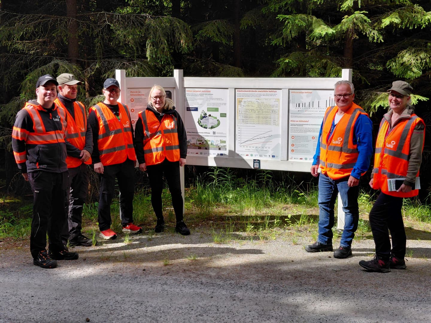 Kuusi huomioliivistä henkilöä maasto-opasteen luona kesällä. 