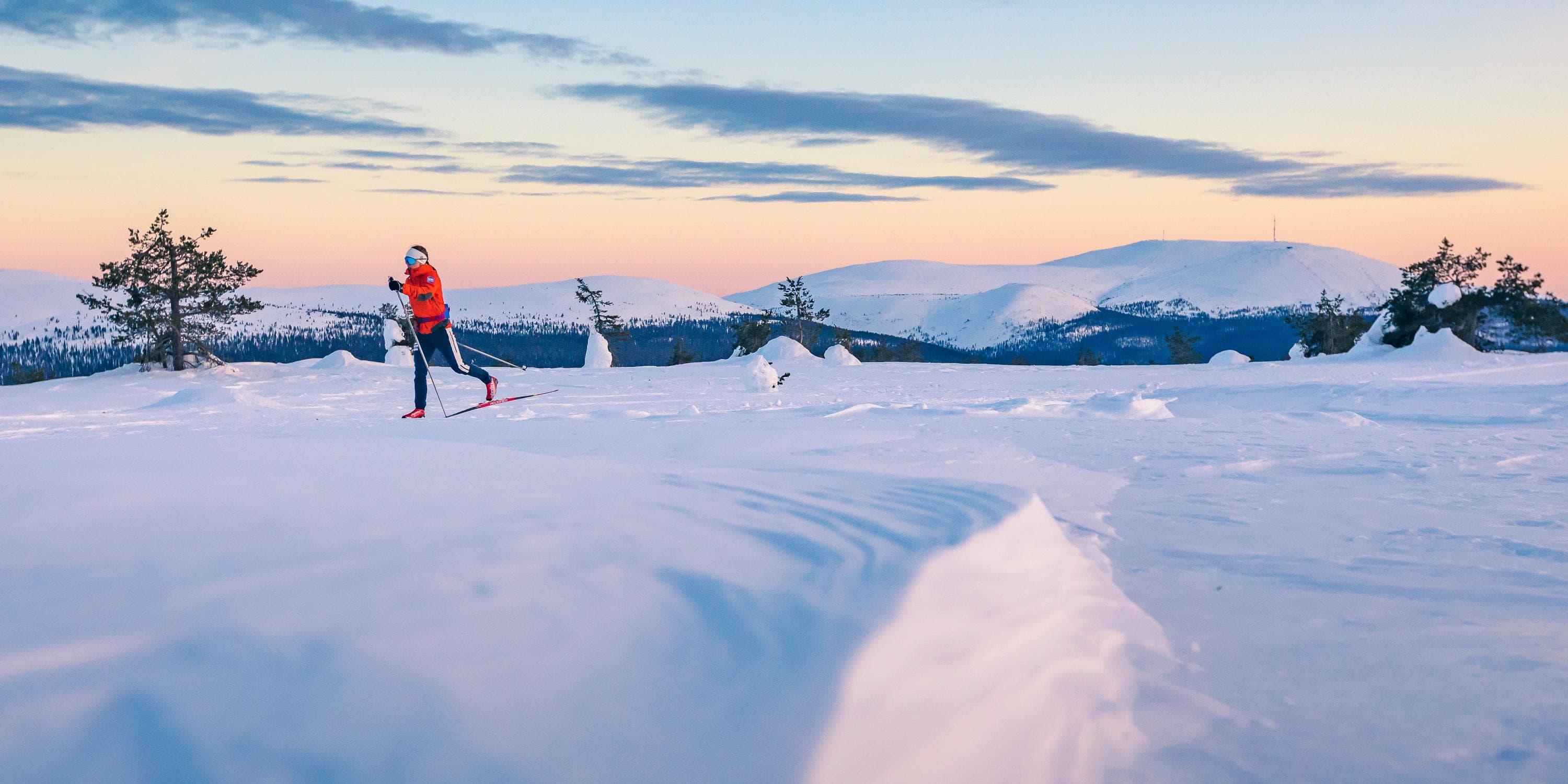 Kuva: Rami Valonen / Metsähallitus Hiihtäjä hiihtää vauhdikkaasti tunturinlaella. Horisontissa näkyy lisää tuntureita. Taivas on värikäs.