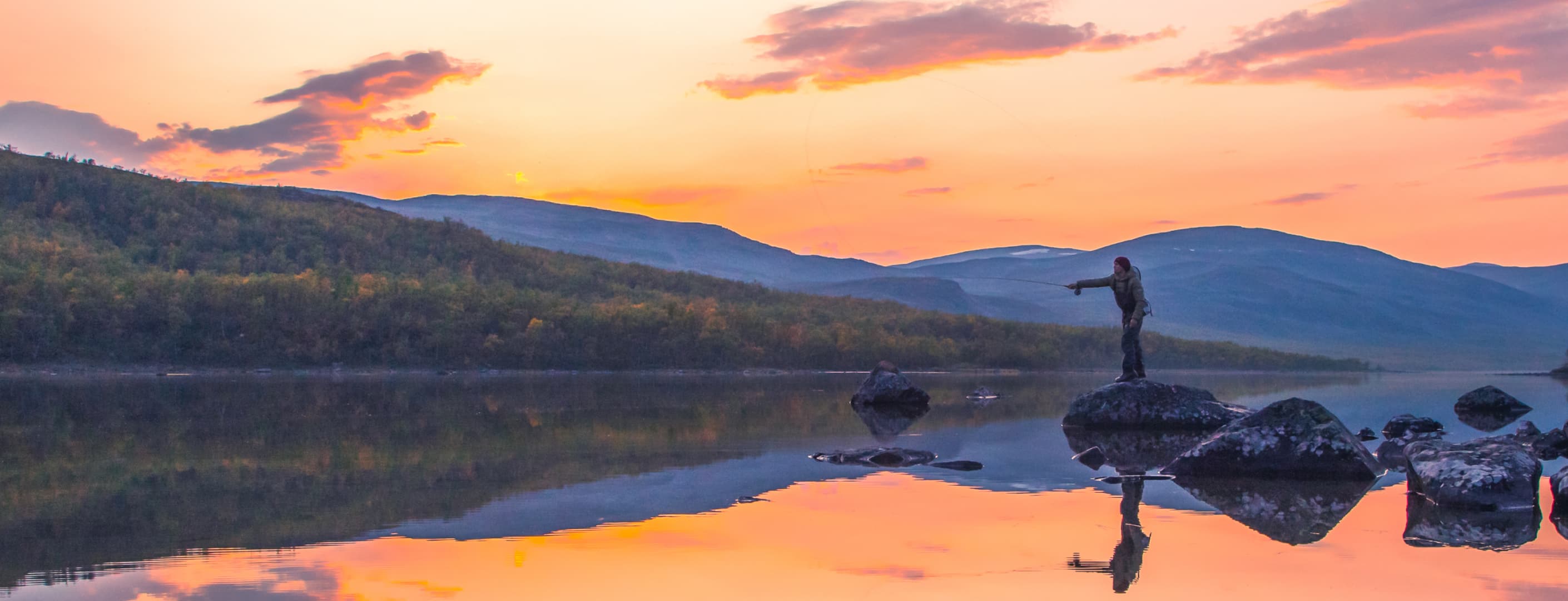 Image: Rami Valonen A fisherman stands on a rock in an autumn wilderness. In the background, there are fells, a sunset, and a calm water surface.