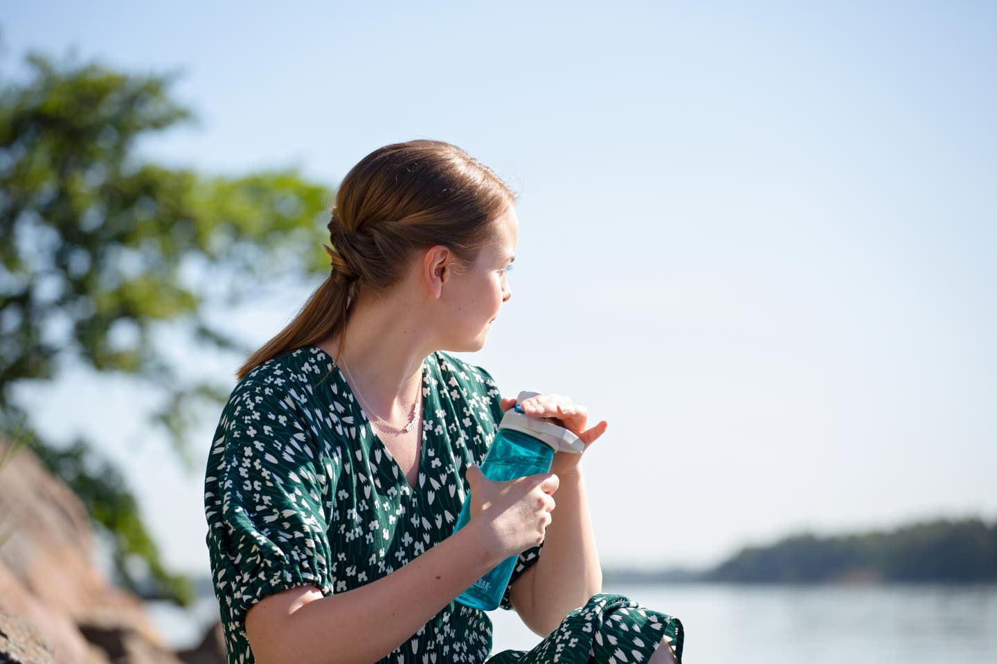 Image: Katri Lehtola / Keksi A person in a green summer dress is sitting on a seaside rock, looking out at the sea away from the camera, with a green water bottle in their hand.