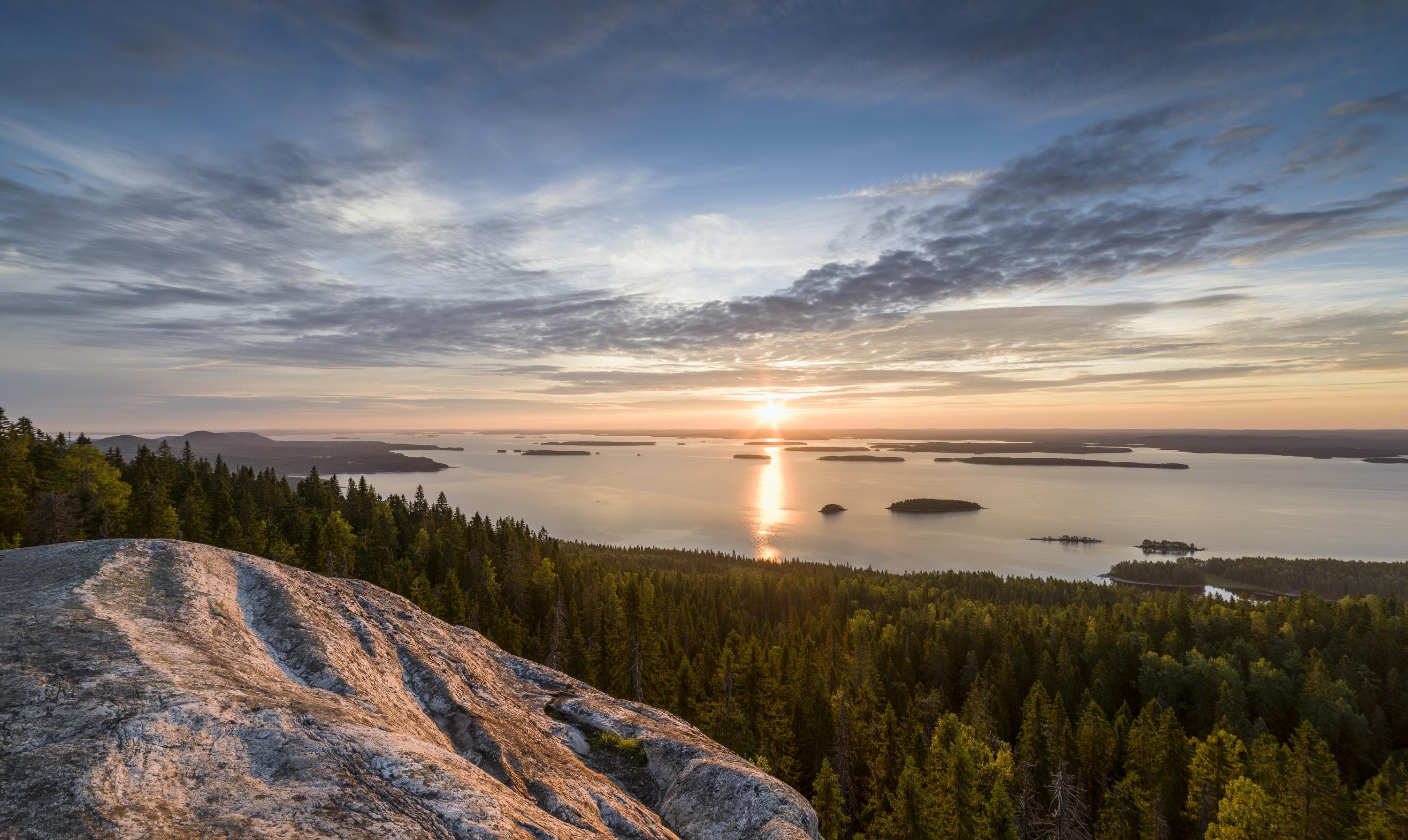 I förgrunden finns det en klippa och skog, med en sjöutsikt i bakgrunden. Solen håller på att gå upp.