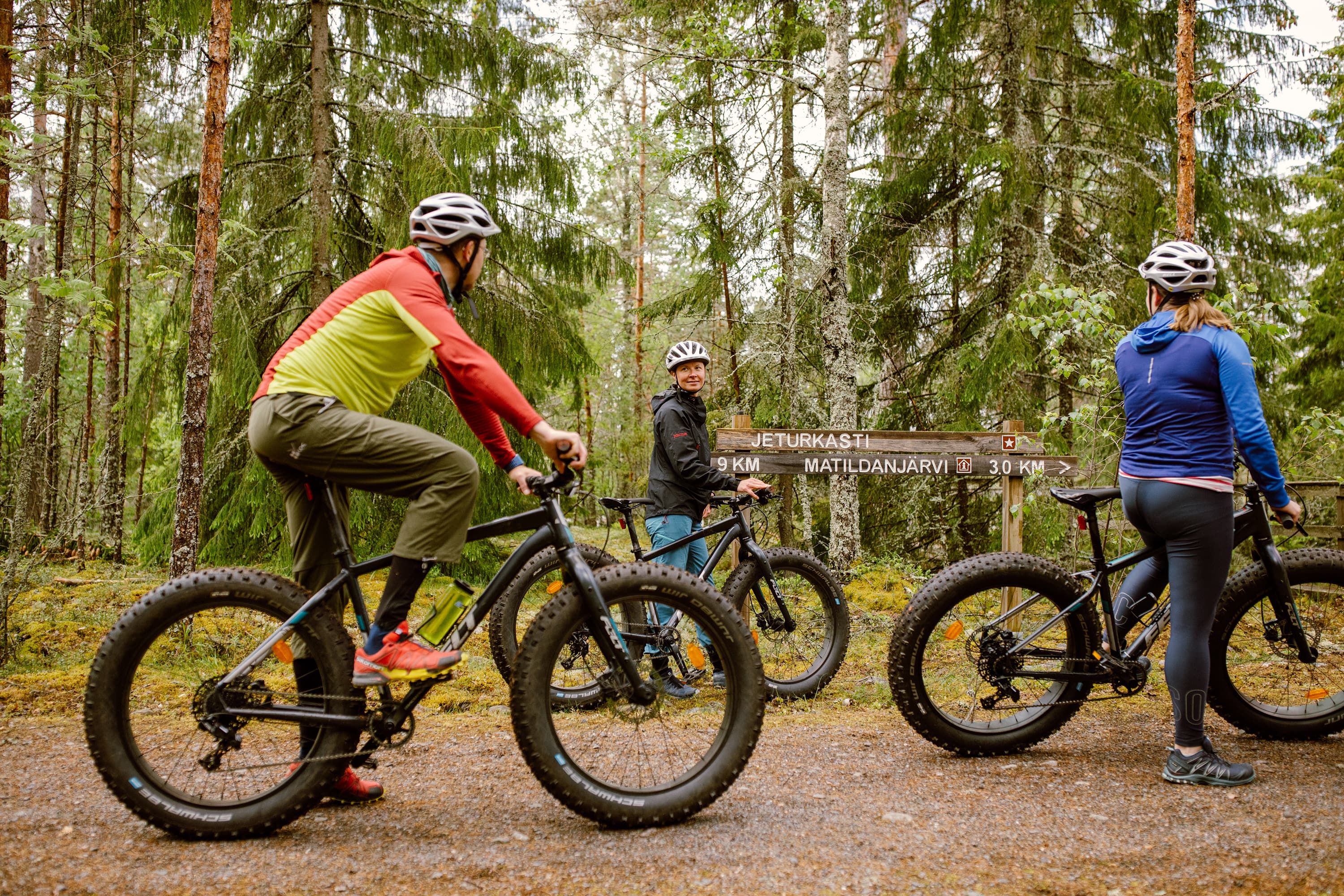 Kuva: Natura Viva Kolme henkilöä ajaa maastopyörillä metsäpolulla. Heillä on yllään pyöräilyvaatteet ja kypärät. Metsä on tiheä ja maassa on pudonneita lehtiä, mikä viittaa syksyyn.