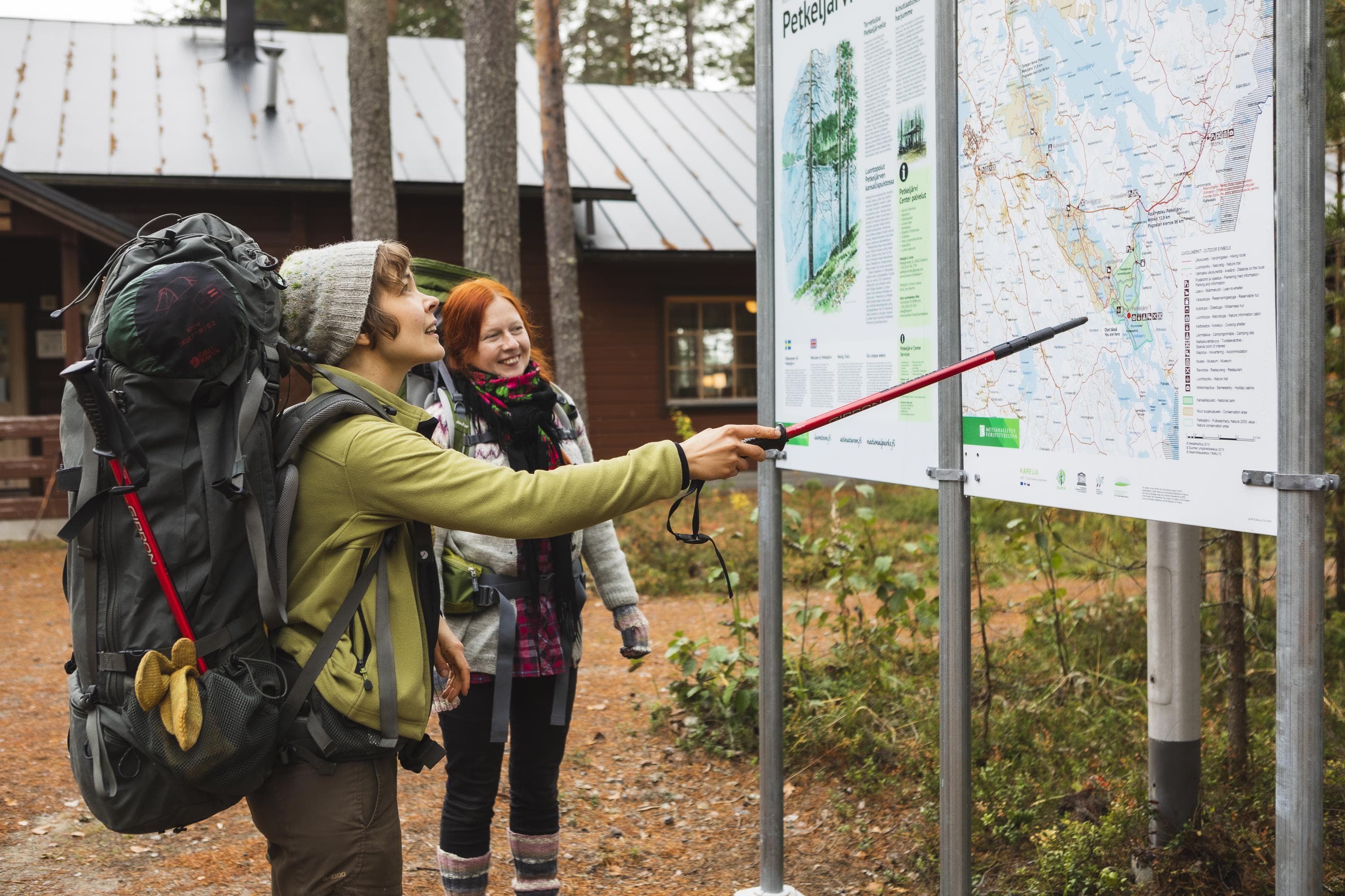 Bild:  Ulla Keituri Två vandrare med ryggsäckar på ryggen står och undersöker en stor informationskarta. I bakgrunden skymtar en träbyggnad.
