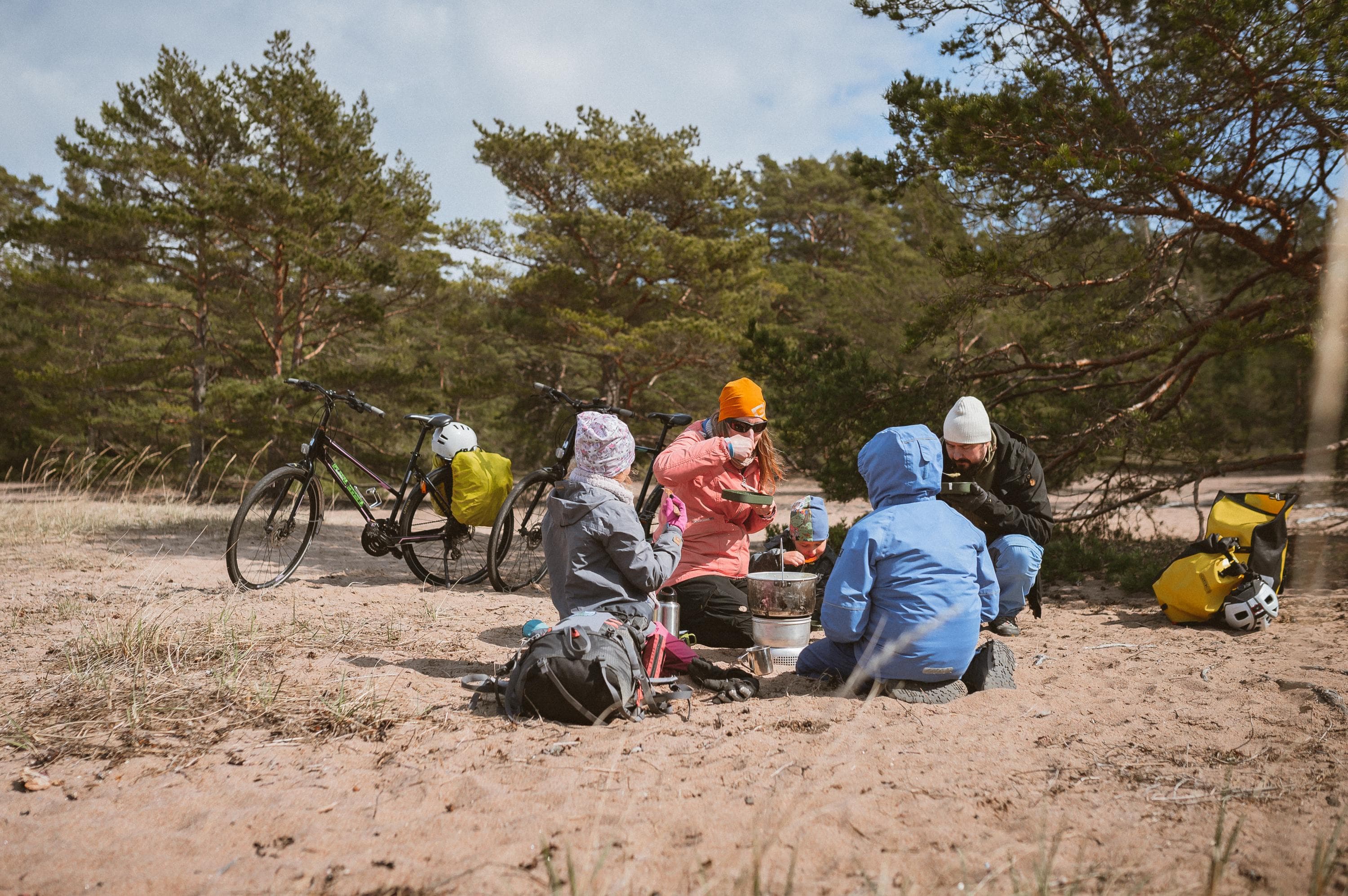 Kaksi aikuista ja kaksi lasta , jotka ovat selin, istuvat hiekalla mäntyjen edessä syömässä. Heillä on retkikeitin keskellään ja takana on kaksi aikuisten pyörää, joissa on pyörälaukut.