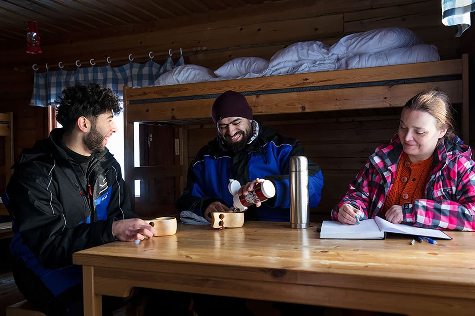 Bild: Kuvatoimisto Keksi. Tre personer sitter runt bordet i stugan.