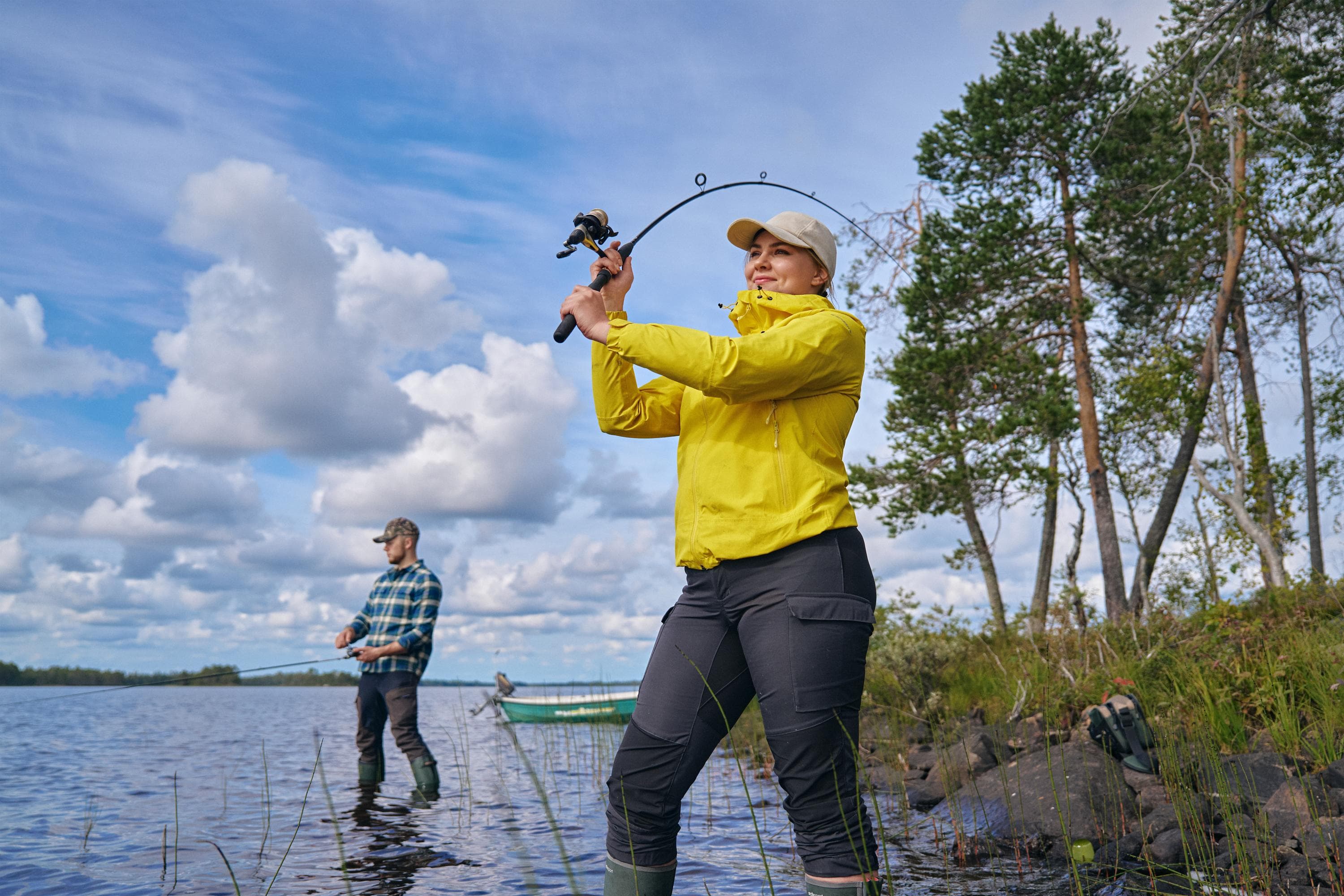 Kaksi vapakalastajaa seisoo rannalla. Kalastaja juuri heittämässä vavalla, taaempana toinen kelailee omaansa.