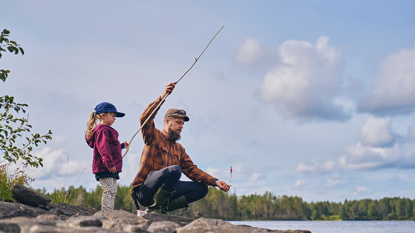 Image: Mikko Törmänen / Keksi A child is standing with a fishing rod in hand, a parent is squatting next to them holding the rod and the bait.