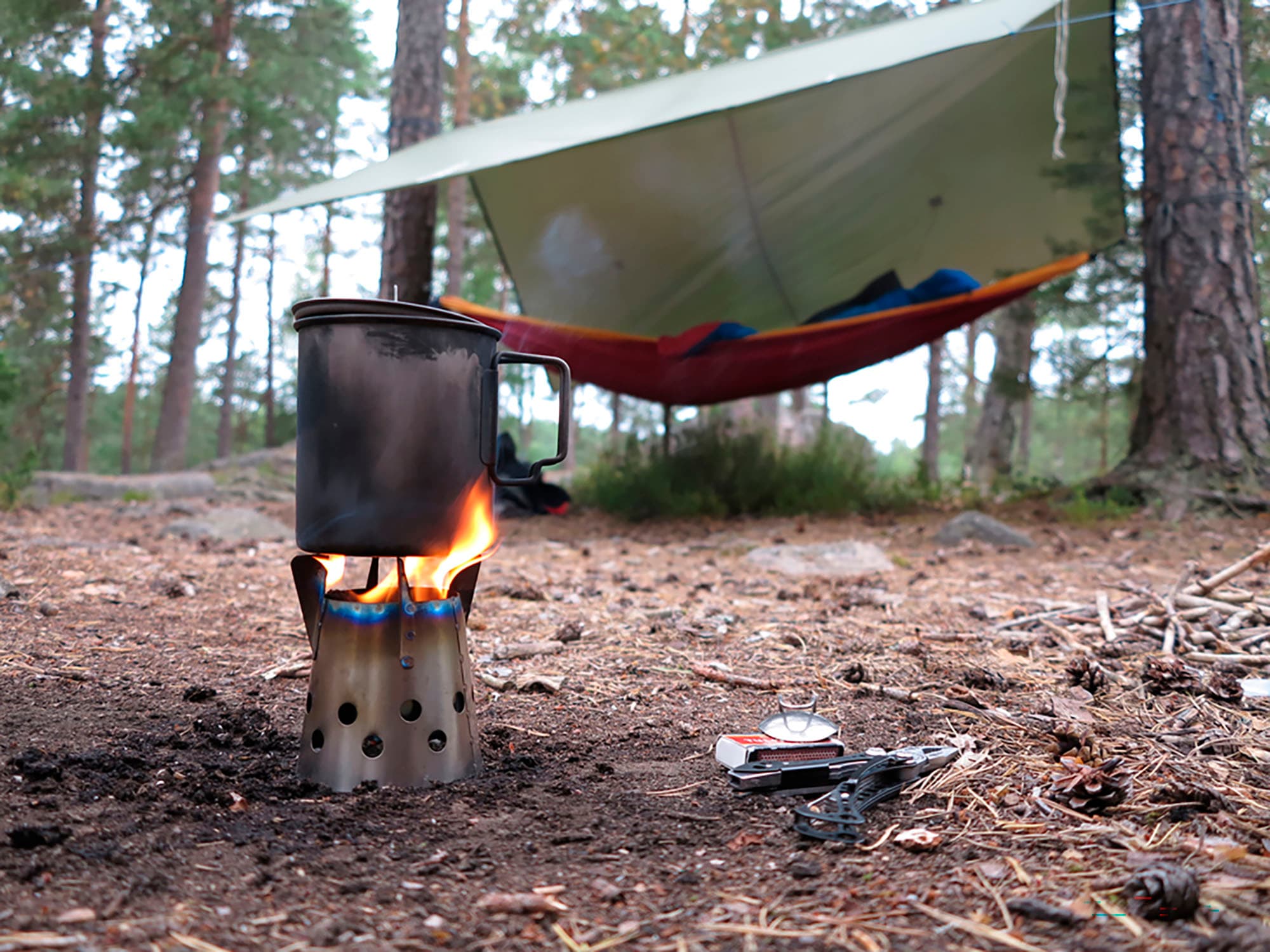 Bild: Suomen Latu / Juhani Metsäpelto I förgrunden en vedspis där maten tillagas. I bakgrunden i skogen hänger en hängmatta mellan två träd och ovanför den finns en presenning.
