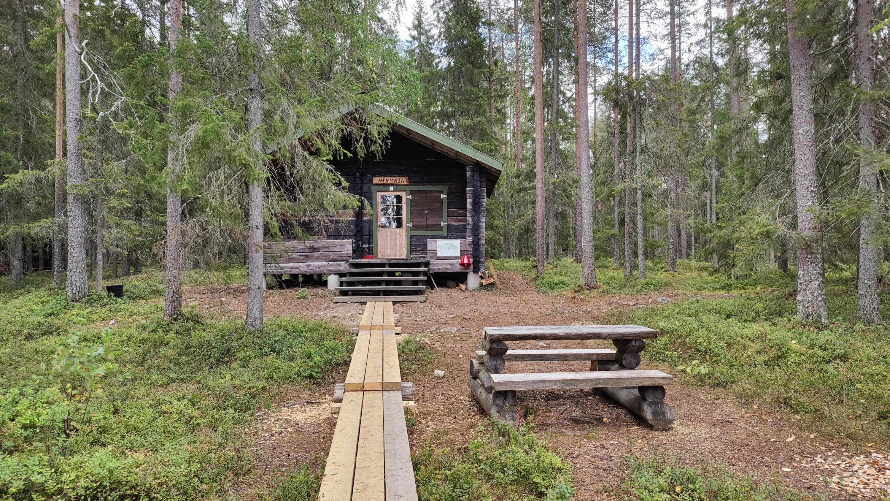 Kuva: Ulla Matturi Pitkokset johtavat hirsituvalle metsän keskellä. Pitkosten oikealla puolella on ruokailupöytä.