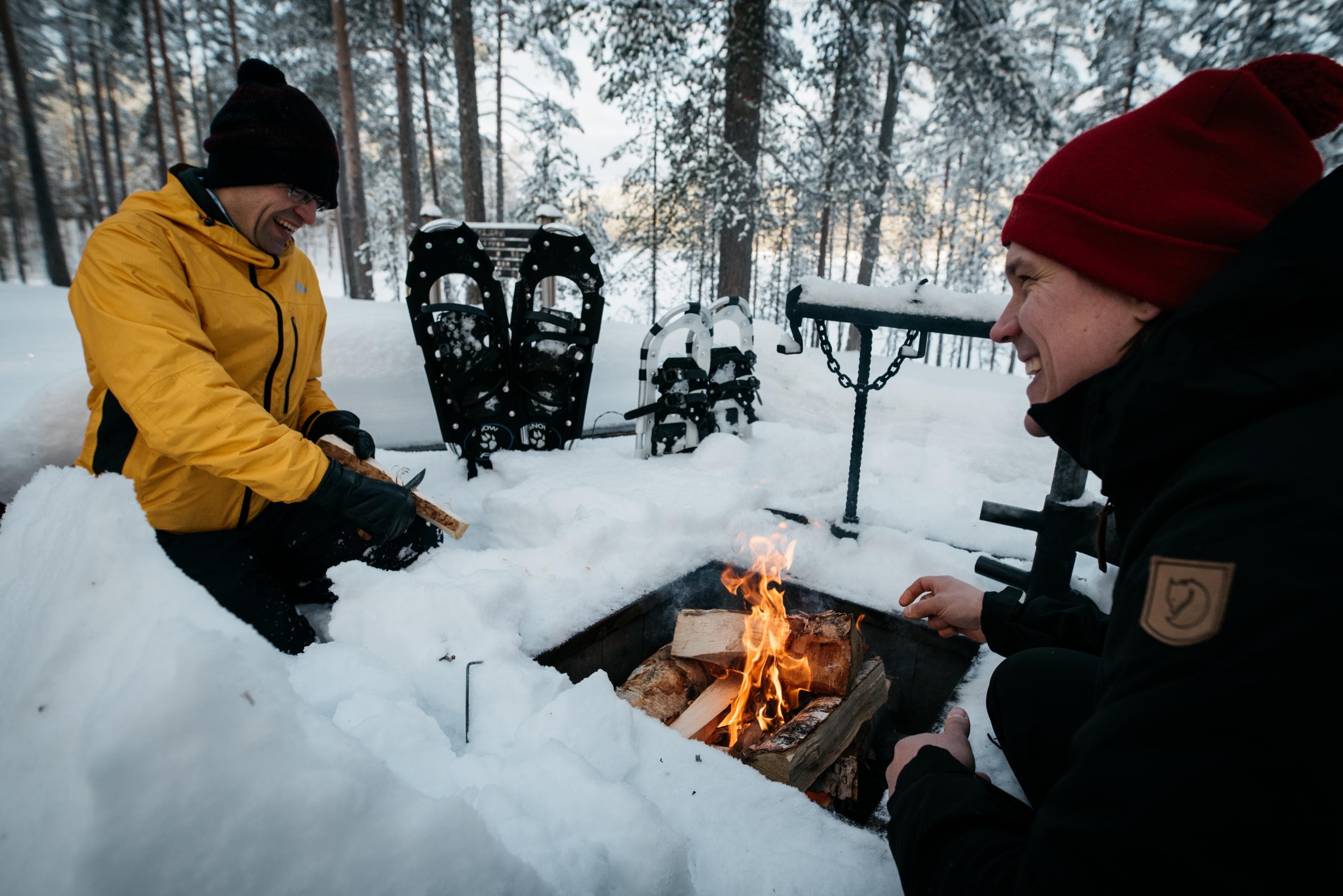 Image: Eeva Mäkinen Two hikers have lit a fire at a snow-covered campfire site. The snowshoes are standing upright in the snow.