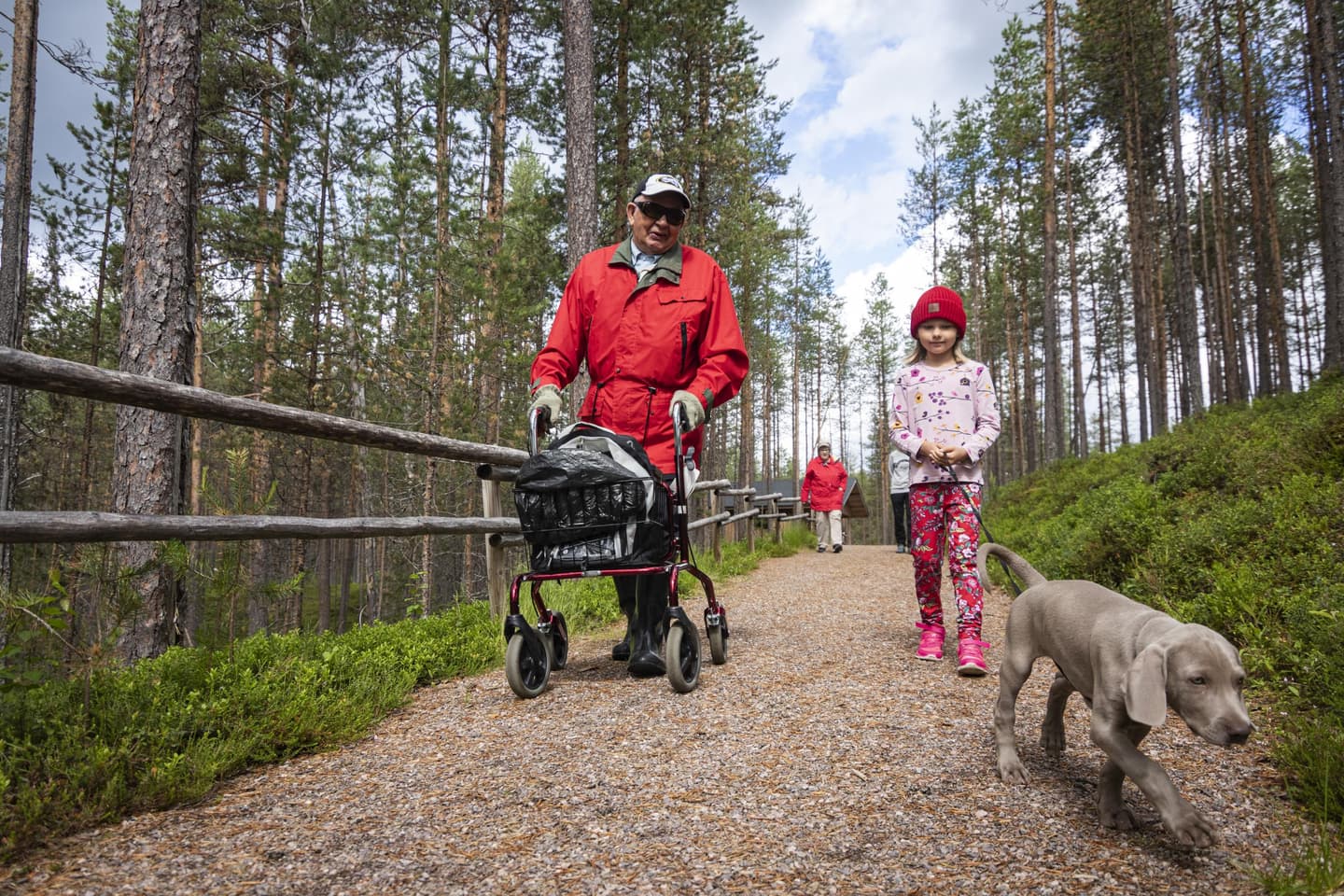 Kuva: Jonna Kalliomäki Vanhempi retkeilijä rollaattorin kanssa ja koiraa taluttava lapsi mäntymetsässä kävelemässä leveää hiekkapolkua kameraa kohti. Polun vieressä kaide.
