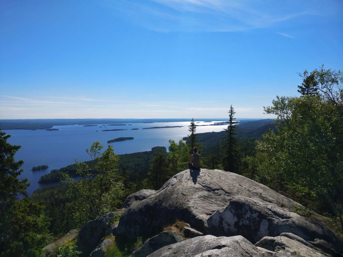 Retkeilijä istuu kesällä korkealla kalliolla. Hän katsoo alhaalla avautuvaa järvimaisemaa.