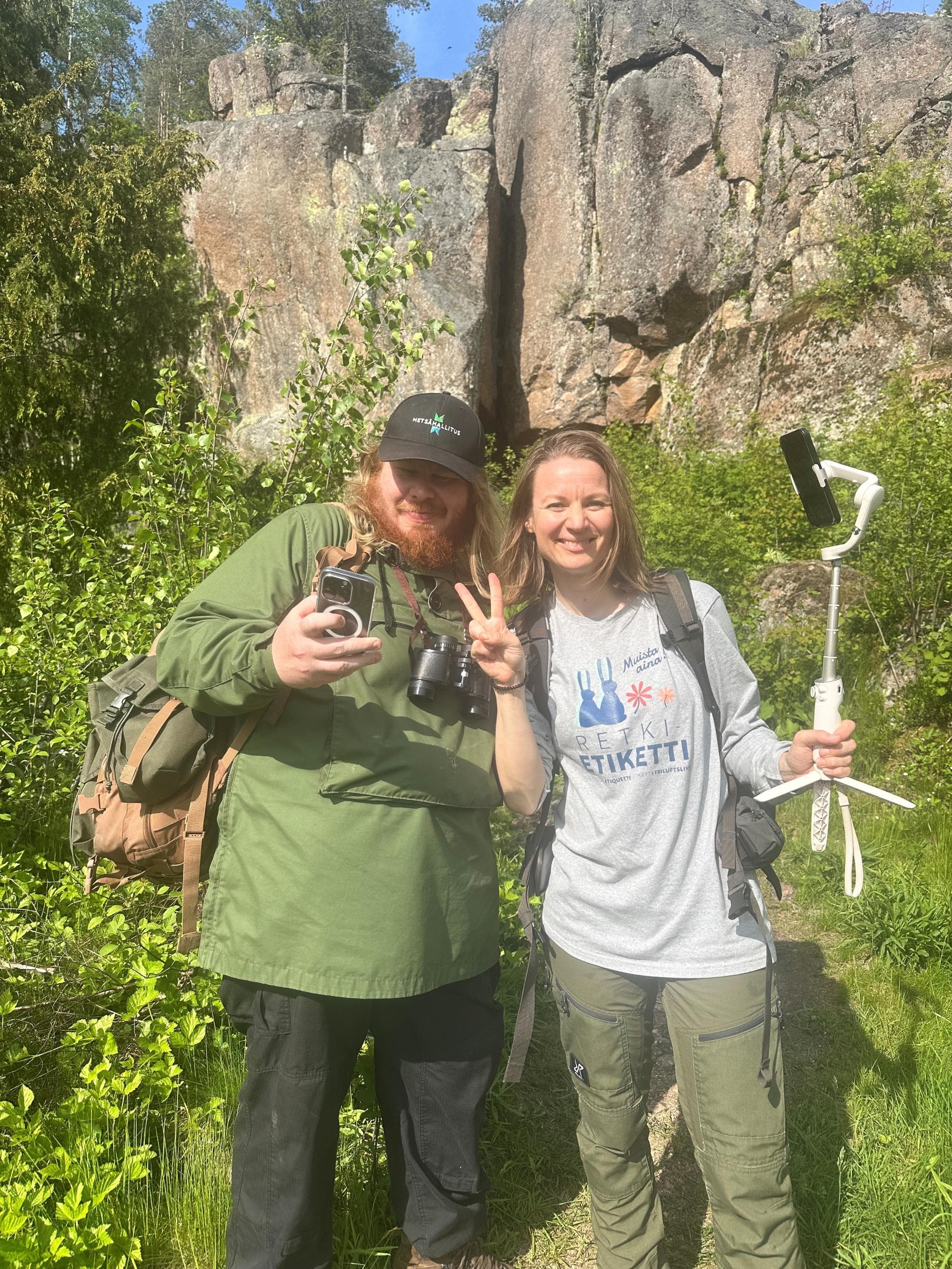 Kuva: Kirsi Kallio Kaksi henkilöä poseeraa kuvaajalle kallioseinämän edessä kesällä. Toisella on kädessään kännykkä ja kaulassa kiikarit ja toisella on kädessään selfiekeppi ja päällään Retkietiketti-paita.