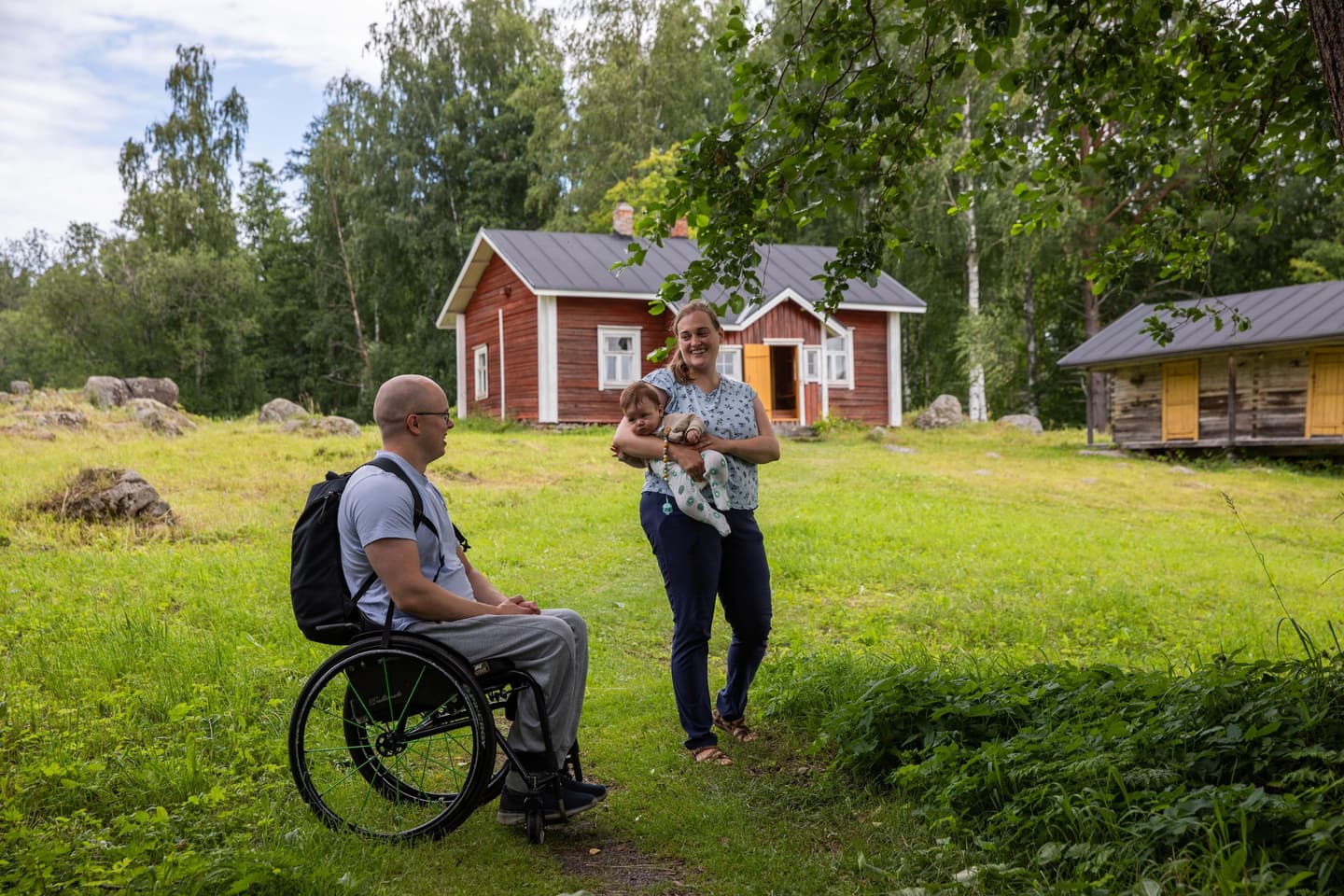 Kuva: Saara Lavi Kaksi retkeilijää katsoo toisiaan hymyillen. Toinen istuu pyörätuolissa ja toisella on sylissään vaiva. Taustalla on puisia rakennuksia.