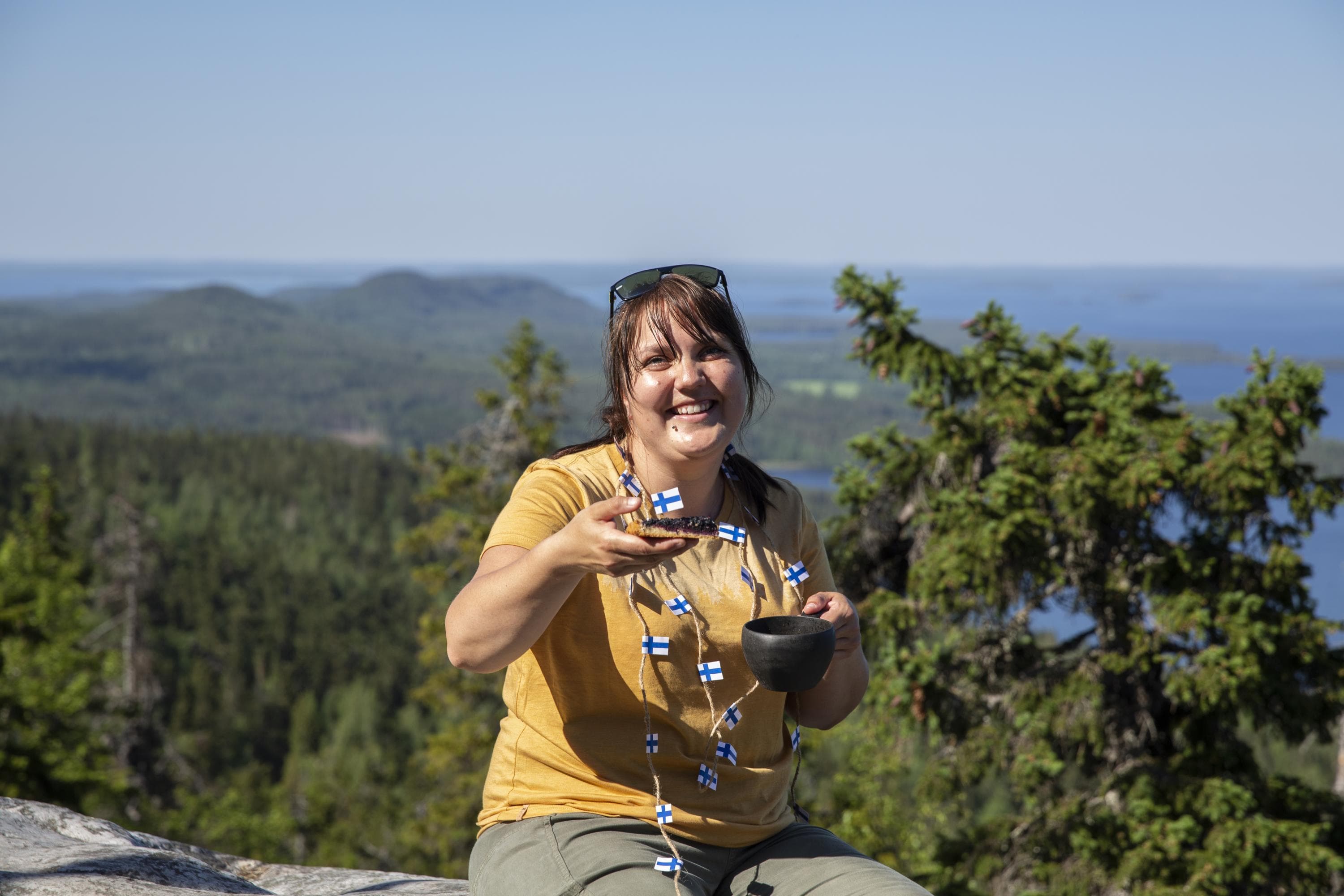Bild: Saara Lavi En vandrare håller en bit blåbärspaj i ena handen och en kuksa i den andra. Runt halsen hänger ett flaggband gjort av små finska flaggor. Vandraren sitter på klippor med utsikt över ett landskap med höjder och vatten.