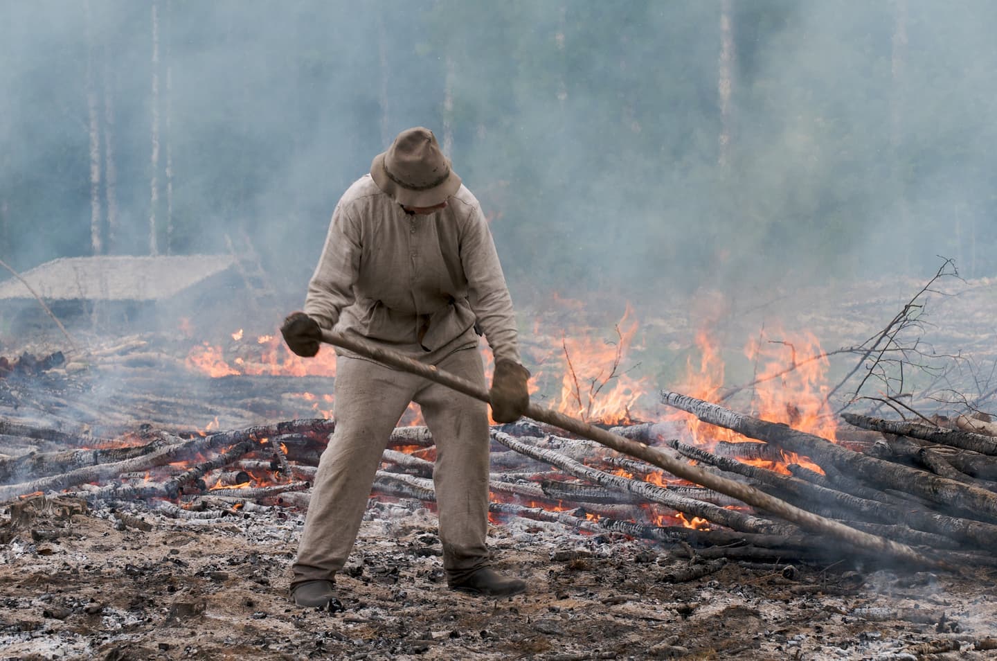 Kuva: Jouni Borgman Perinneasuinen viertää palavaa kaskea. Savun läpi siintää taustalla oleva rakennus ja metsänreuna.