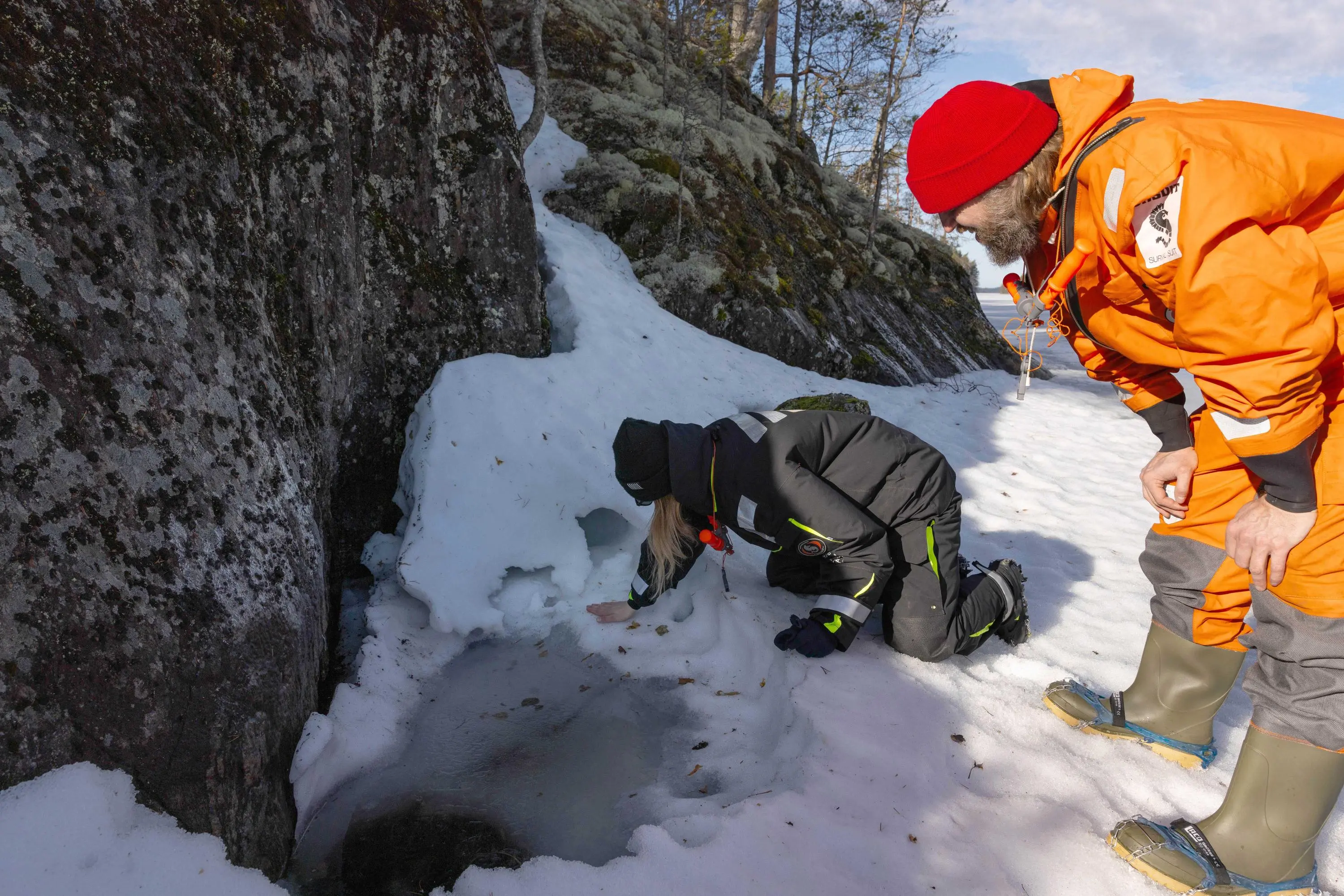 Kuva: Samuel Bloch Kaksi henkilöä on kumartuneena rantakallion vieressä olevan osittain romahtaneen saimaannorpan pesäluolan äärellä.
