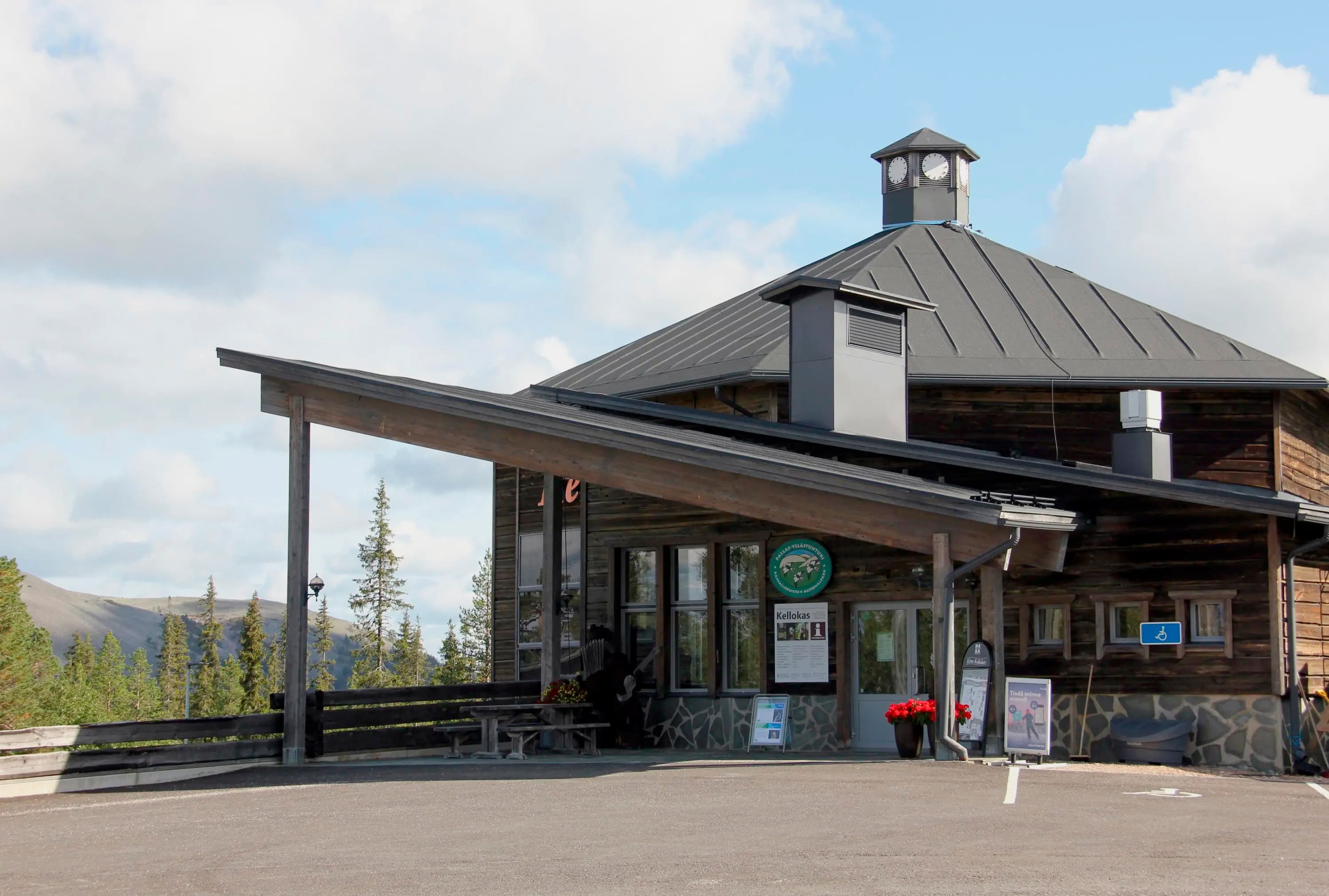 Kuva: Roosa Pyy Hirsisen rakennuksen sisäänkäynti. Rakennuksen katolla on pieni kellotorni ja rakennuksen takana on tunturimaisema.