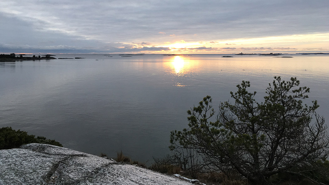 Merenrantaa, merta ja saaria kauempana. Pääosin pilvisellä taivaalla aurinko pilkottaa matalalla.